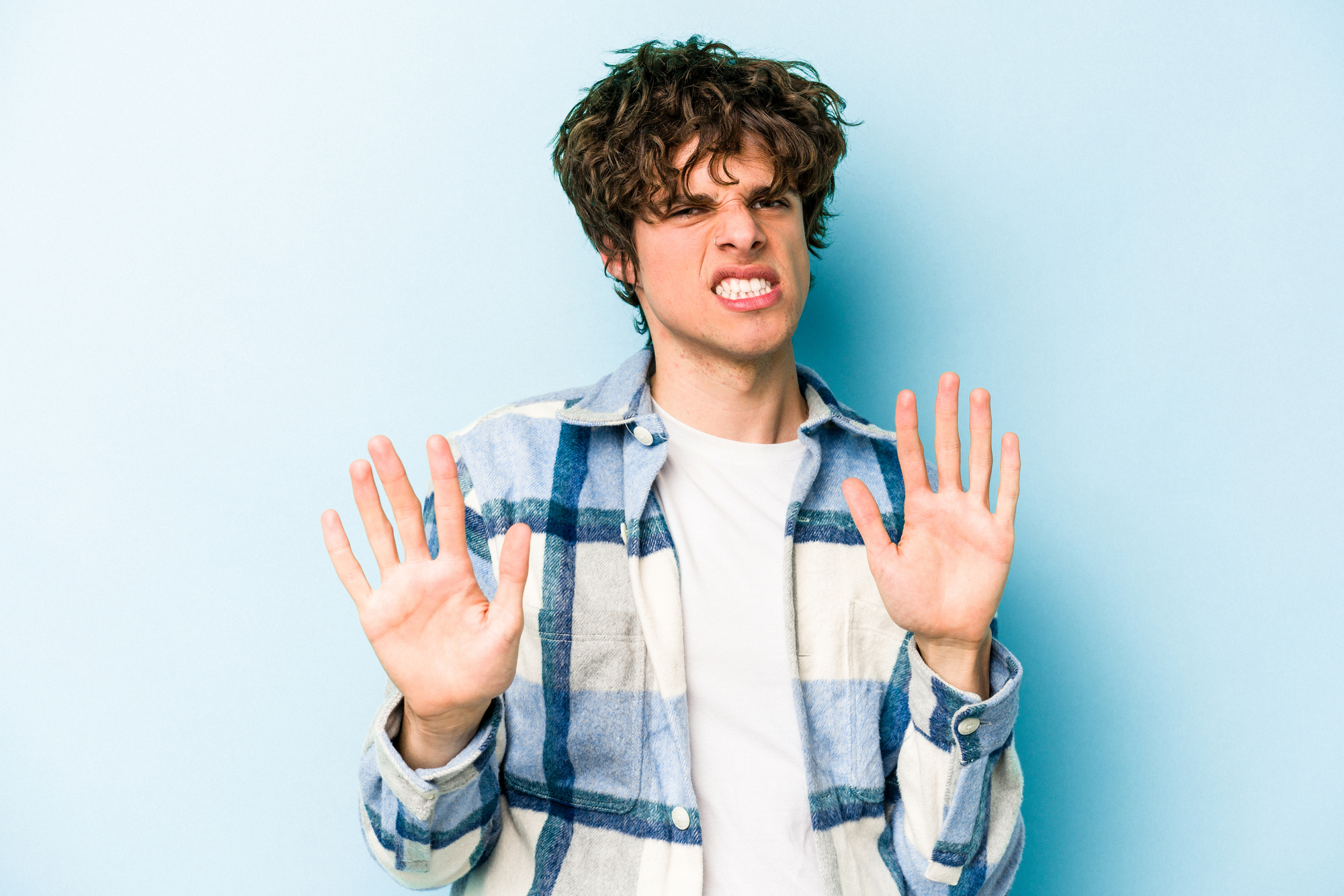 A young man with curly hair and a plaid shirt stands against a light blue background. He has a playful, exaggerated expression with his mouth slightly open and palms facing forward as if playfully saying, "No way!.