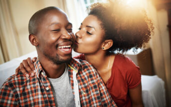 A woman with curly hair lovingly kisses a smiling man on the cheek. They are sitting indoors, and the man is wearing a plaid shirt over a gray t-shirt. The mood is warm and affectionate.