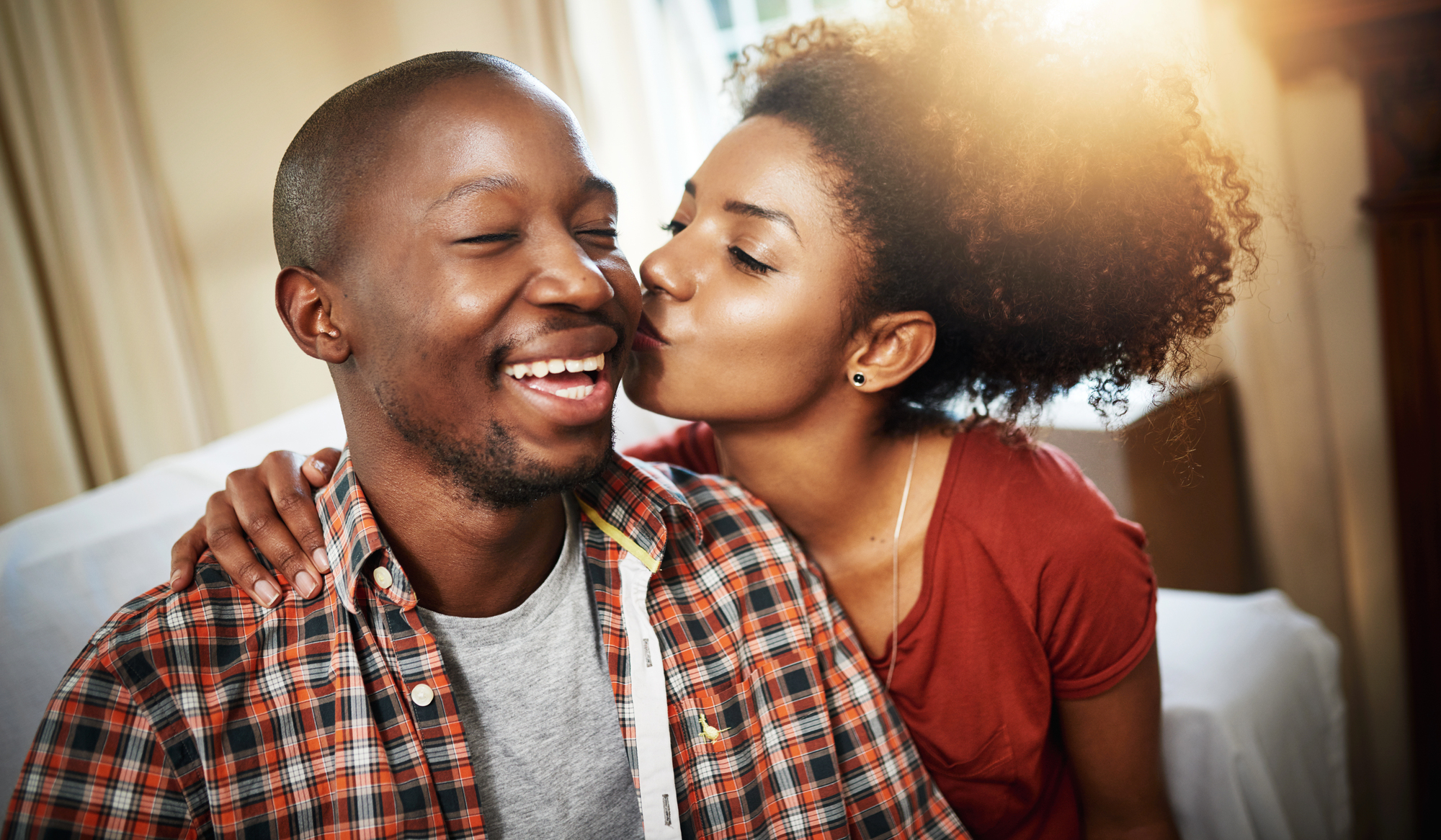 A woman with curly hair lovingly kisses a smiling man on the cheek. They are sitting indoors, and the man is wearing a plaid shirt over a gray t-shirt. The mood is warm and affectionate.