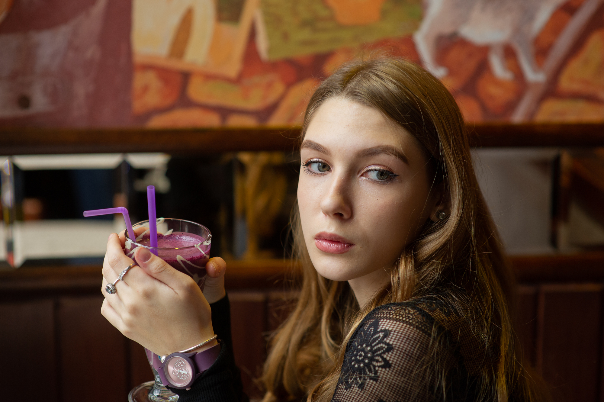 A person with long hair holds a purple drink with two straws, sitting in a cafe. The background features a colorful mural, and the person is wearing a watch and a lace top.