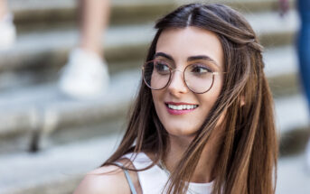 A woman with long brown hair and round glasses smiles while looking away. She is standing on steps, and her hair is gently blowing in the breeze. She is wearing a white top.