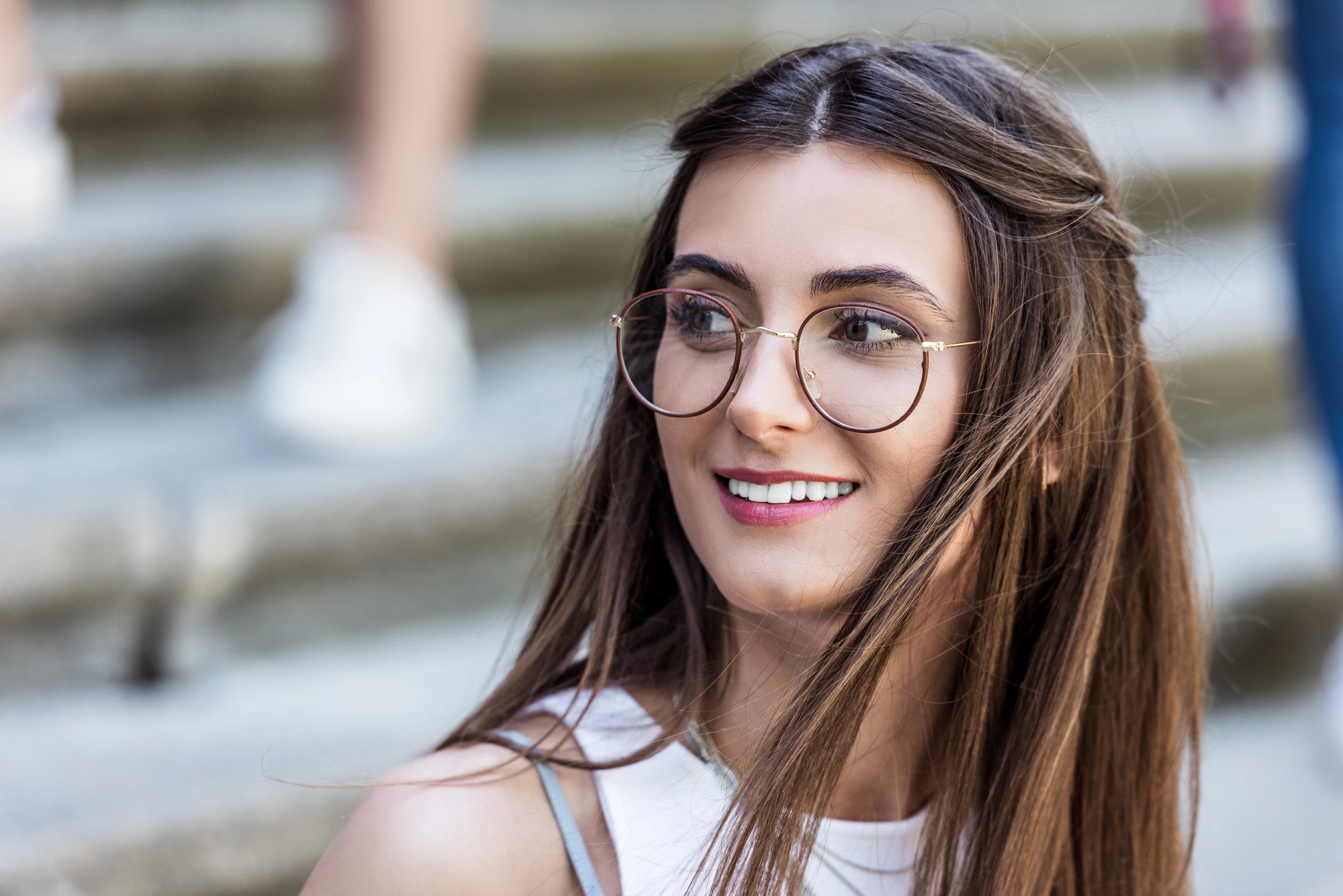 A woman with long brown hair and round glasses smiles while looking away. She is standing on steps, and her hair is gently blowing in the breeze. She is wearing a white top.