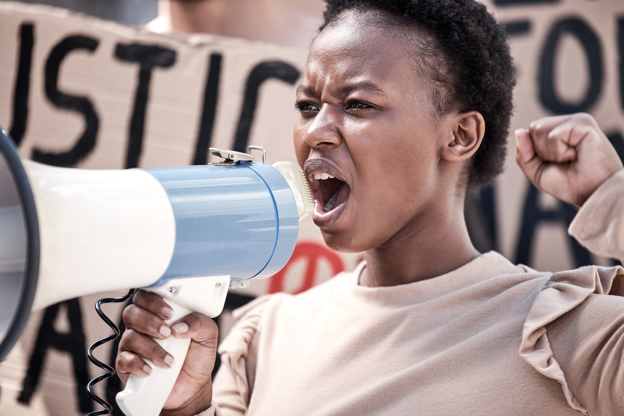 Person passionately speaking into a megaphone at a protest, with a raised fist and a sign with partially visible text in the background.