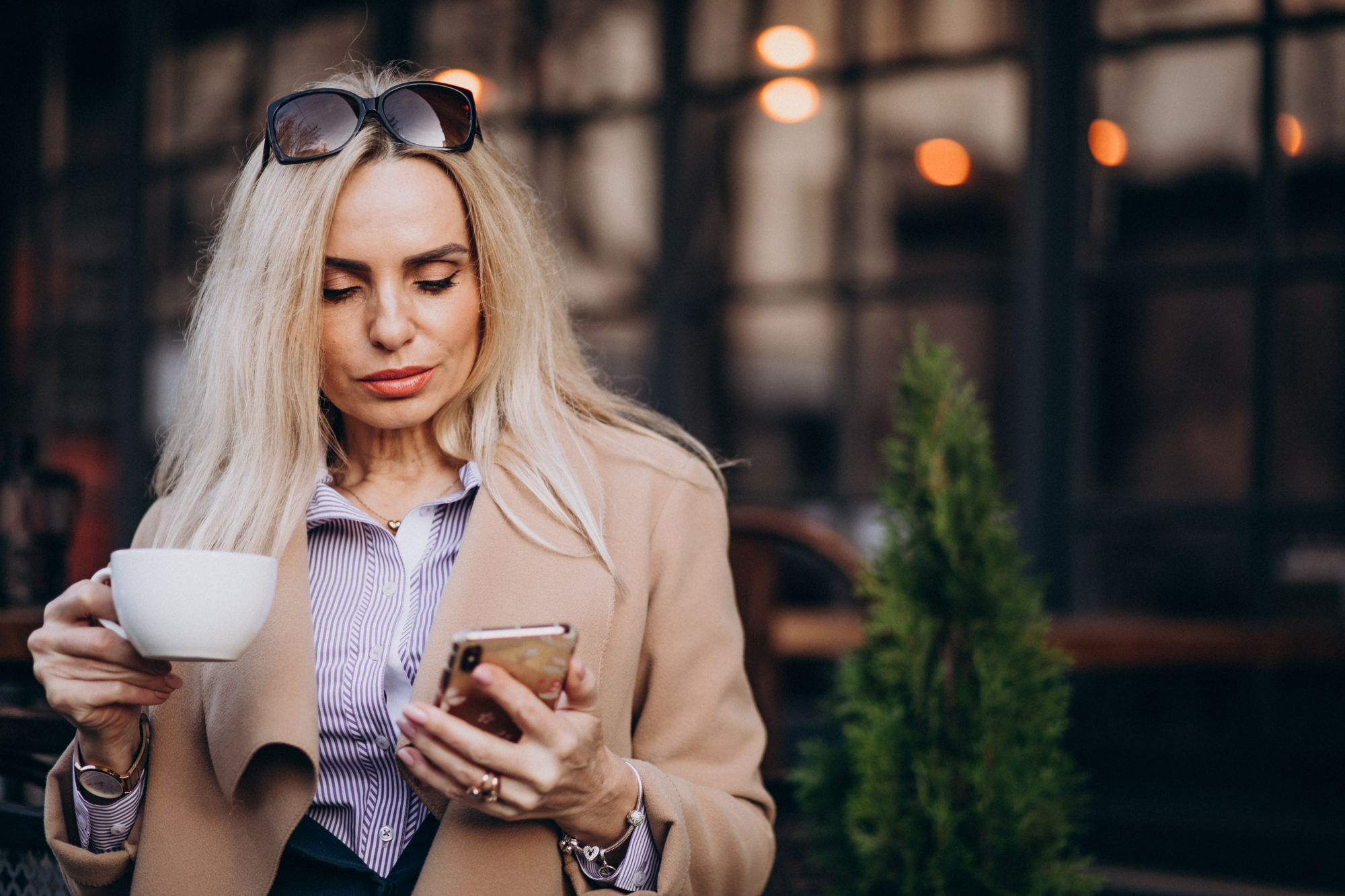 A woman with long blonde hair is sitting outside a café, holding a white coffee cup in one hand and looking at her smartphone in the other. She is wearing sunglasses on her head, a striped shirt, and a beige coat.