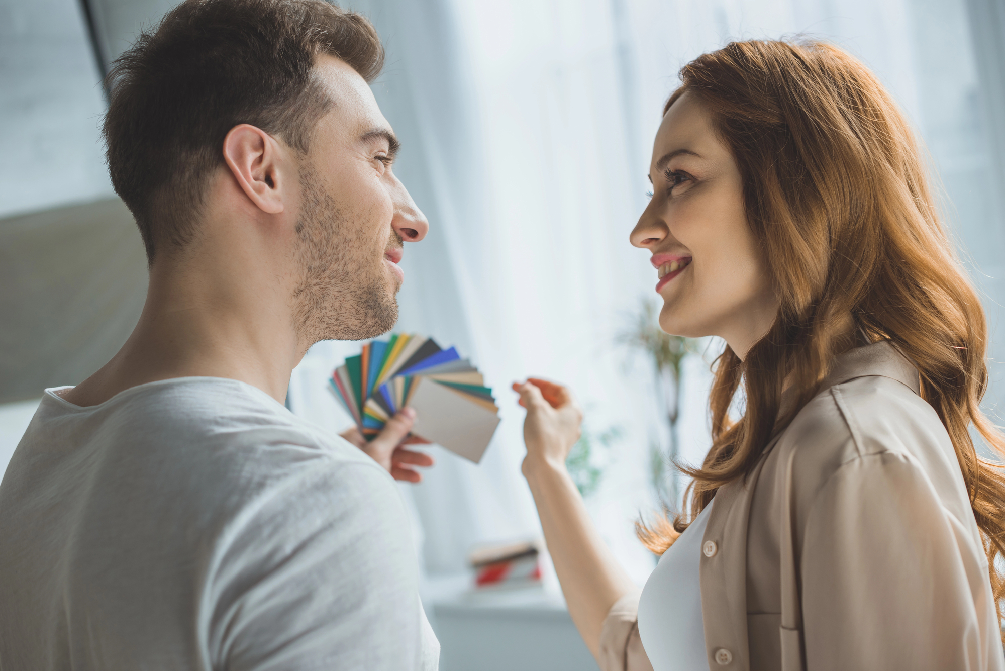 A woman holding a set of colorful paint swatches smiles at a man, who is also smiling. They stand in a bright room, perhaps discussing color choices for decorating. Light filters through sheer curtains in the background.