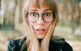 A surprised woman with long hair and glasses holds her hands to her cheeks while standing outdoors in a sunlit wooded area.