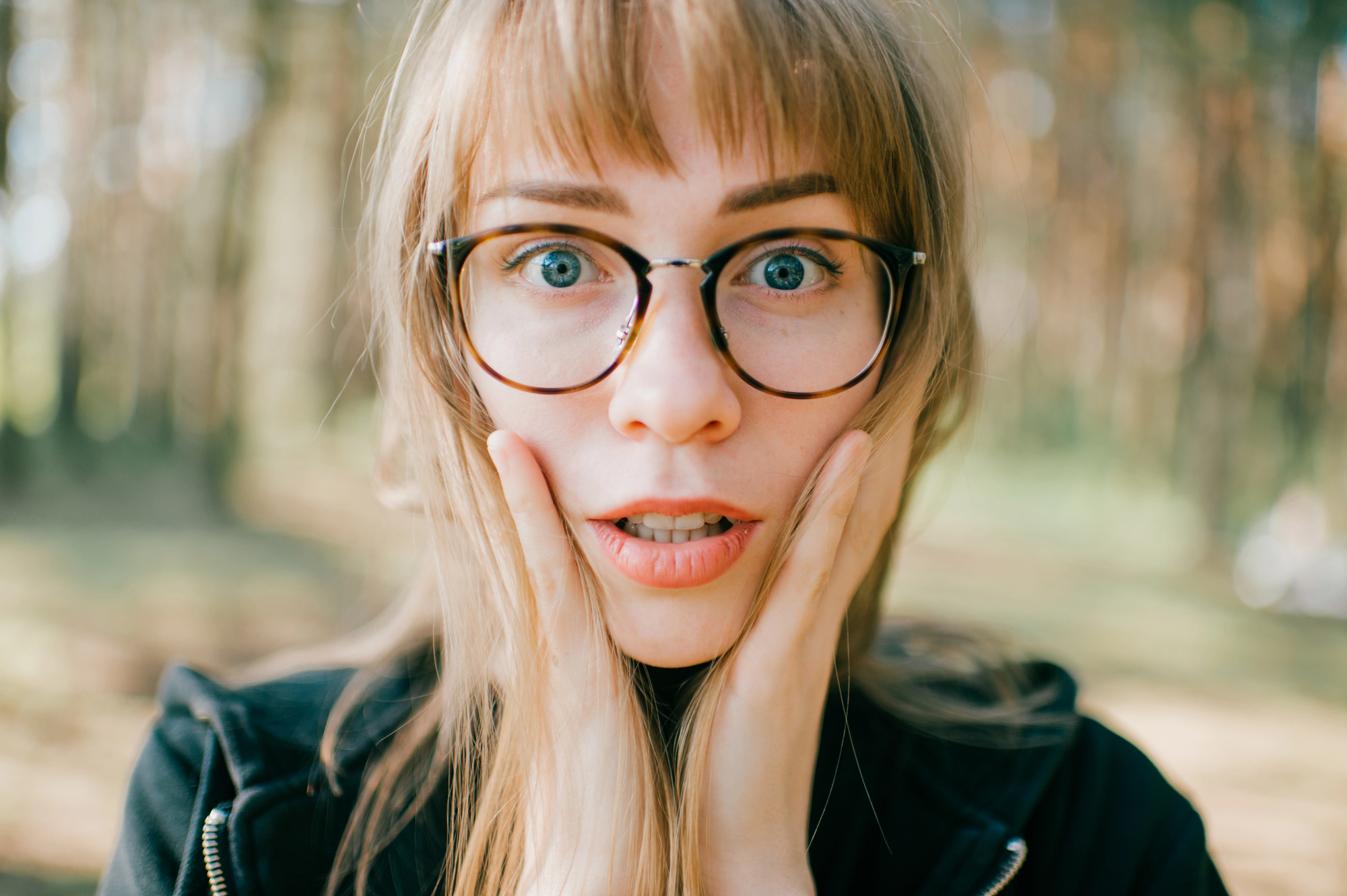 A surprised woman with long hair and glasses holds her hands to her cheeks while standing outdoors in a sunlit wooded area.