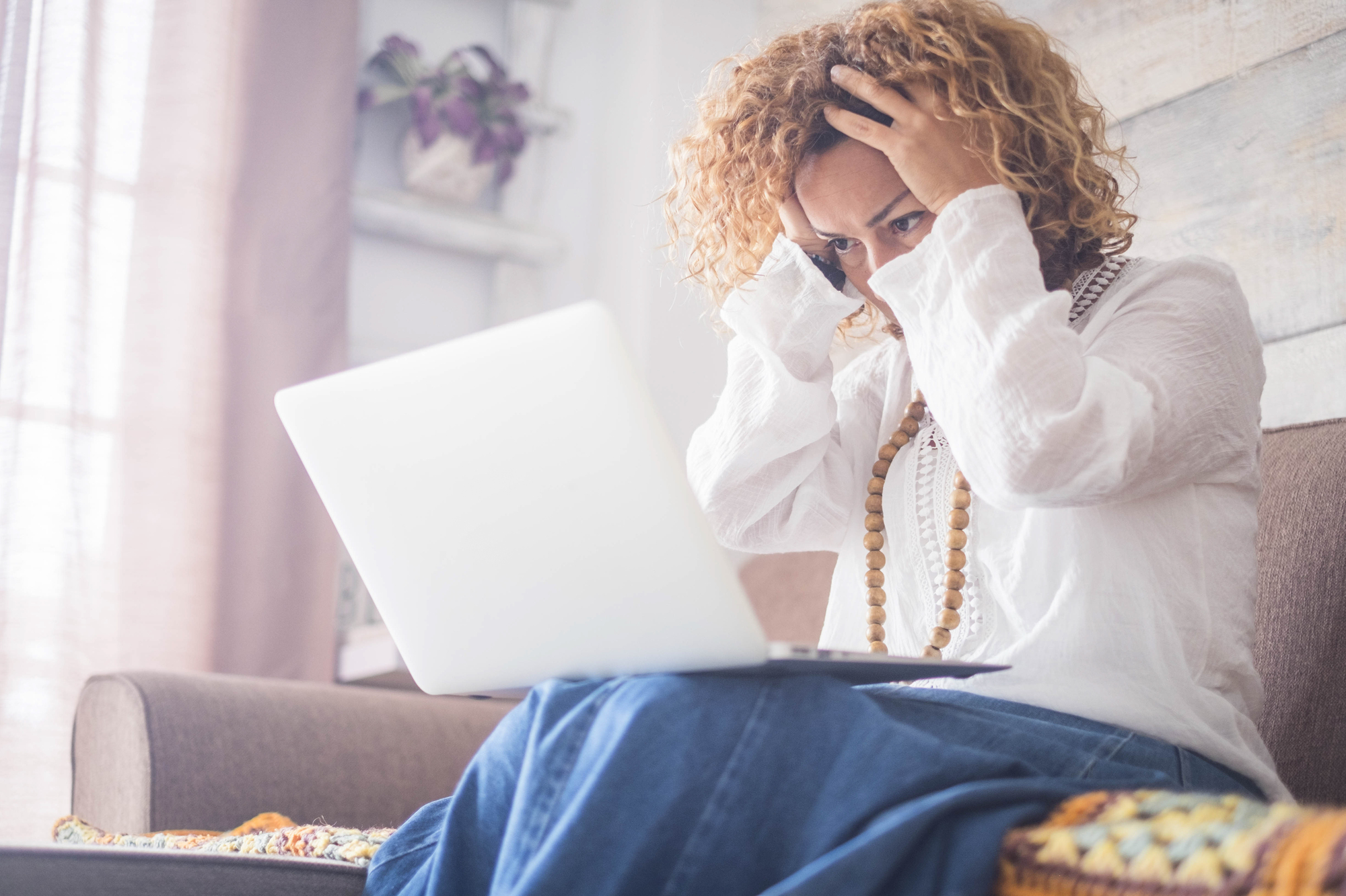 A woman sits on a couch, holding her head in frustration while looking at a laptop. She wears a white shirt and a necklace, and light from a window filters through sheer curtains in the background.