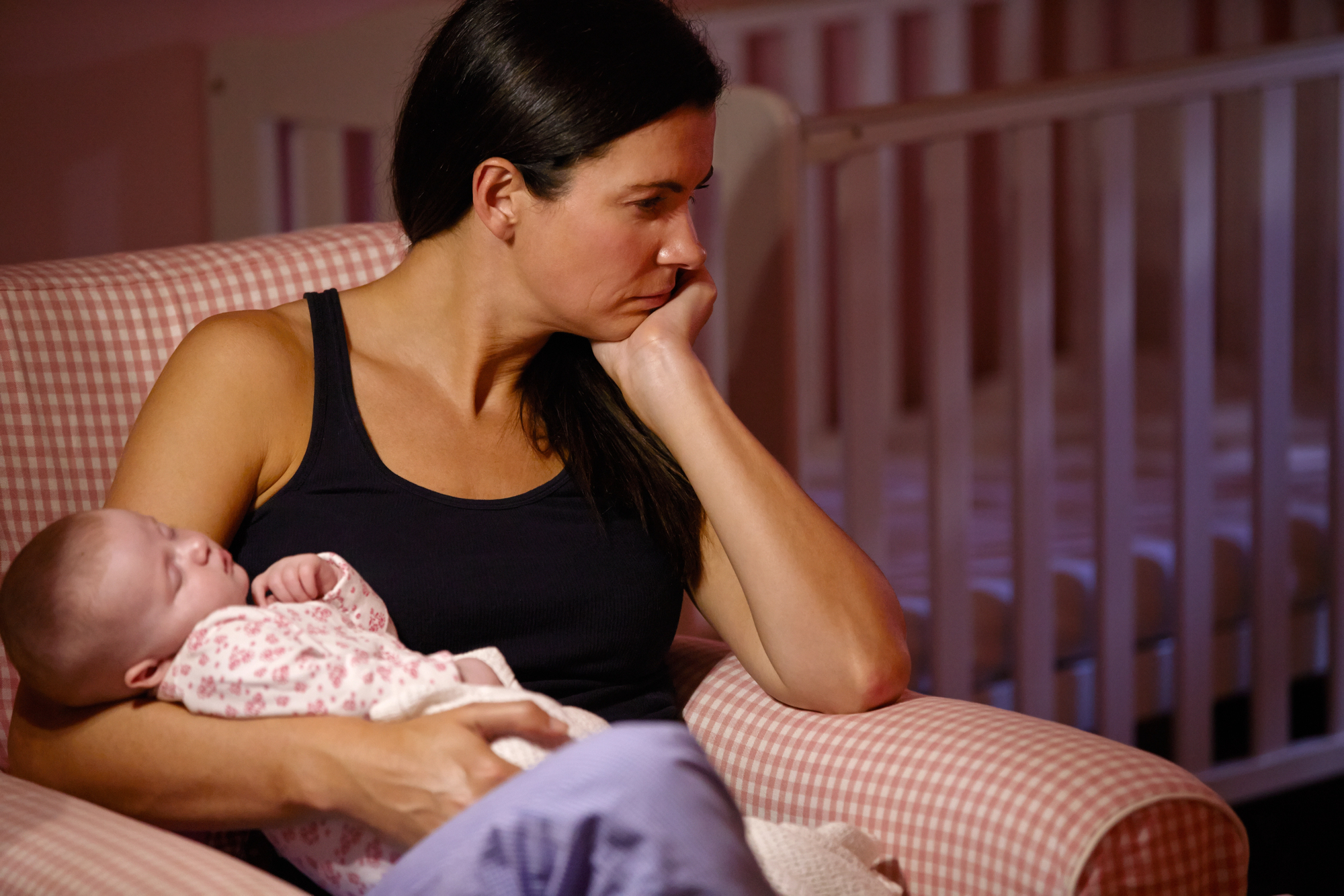 A woman sits in a checkered armchair, resting her head on her hand in thought. She holds a sleeping baby swaddled in a floral blanket. Behind her, a crib is visible, suggesting a cozy, quiet nursery setting.