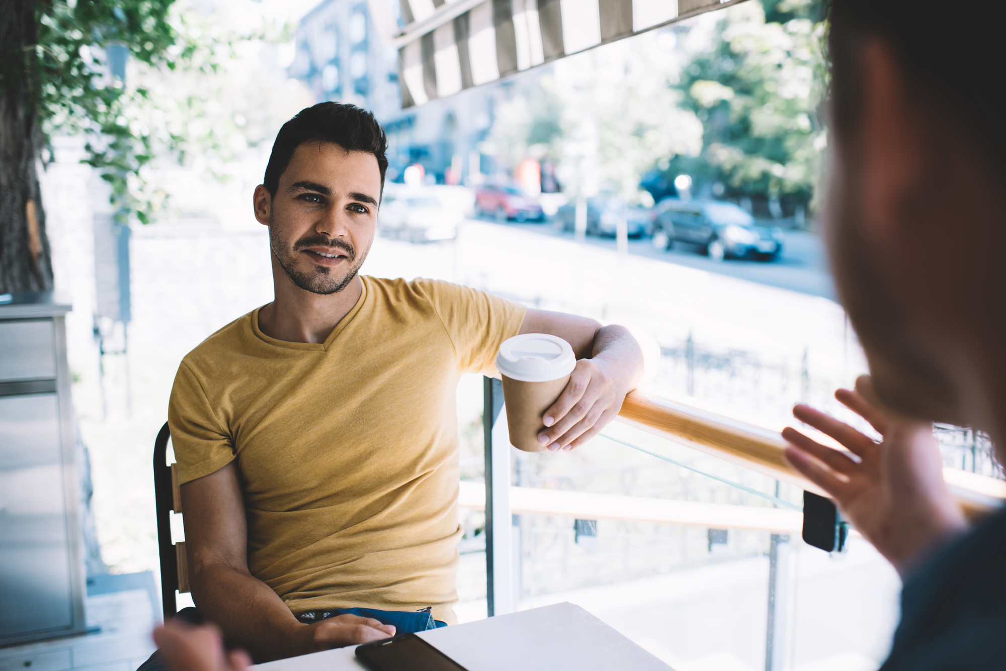 A man in a yellow shirt holds a coffee cup while sitting at an outdoor café table. He is engaging in a conversation with another person who is partially visible in the foreground. The background shows a street scene with trees and buildings.