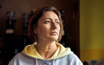 A woman with short brown hair and an earring looks thoughtfully to the side, wearing a hoodie. The lighting creates a warm ambiance, with soft focus on the background.