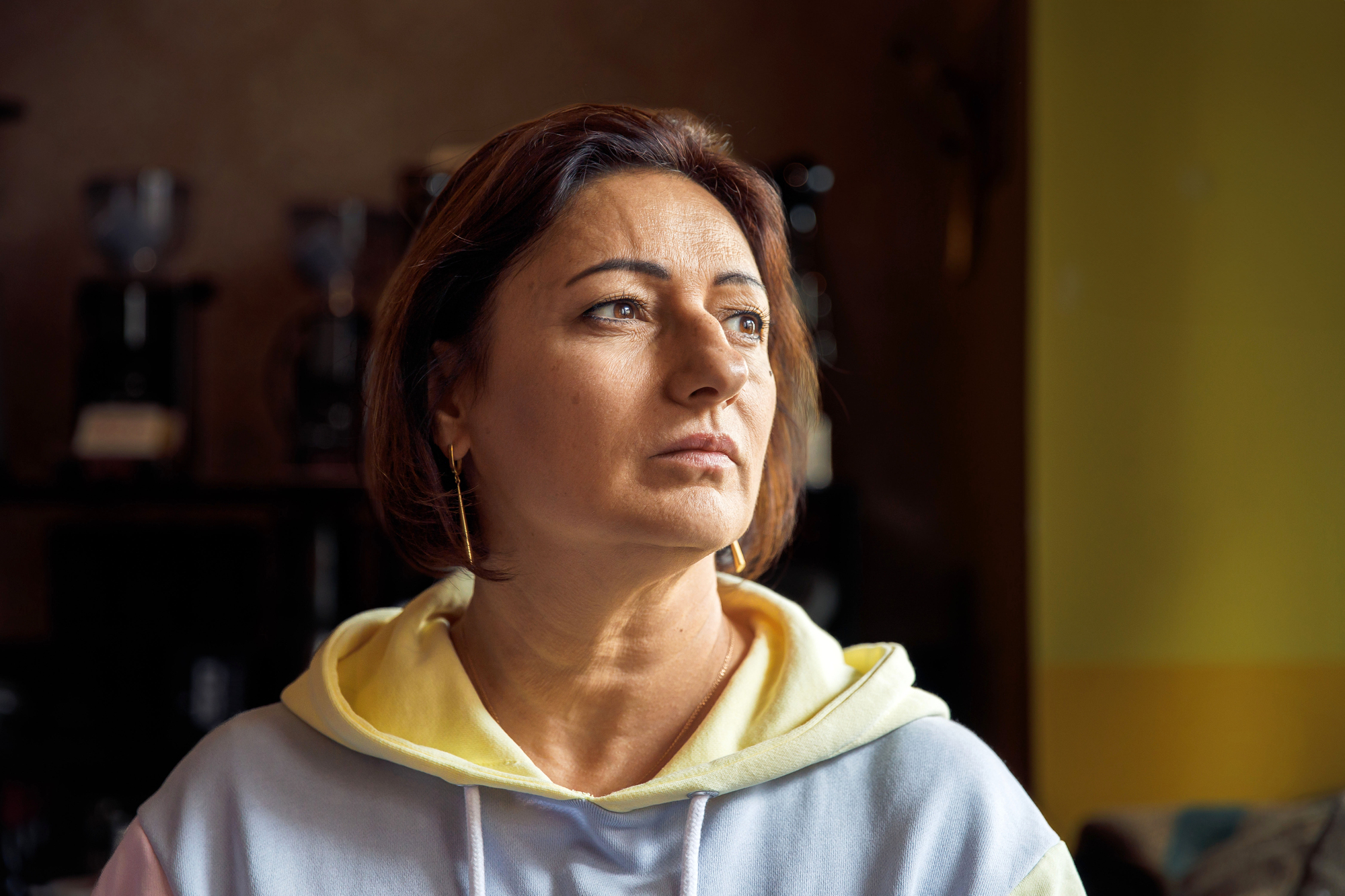 A woman with short brown hair and an earring looks thoughtfully to the side, wearing a hoodie. The lighting creates a warm ambiance, with soft focus on the background.