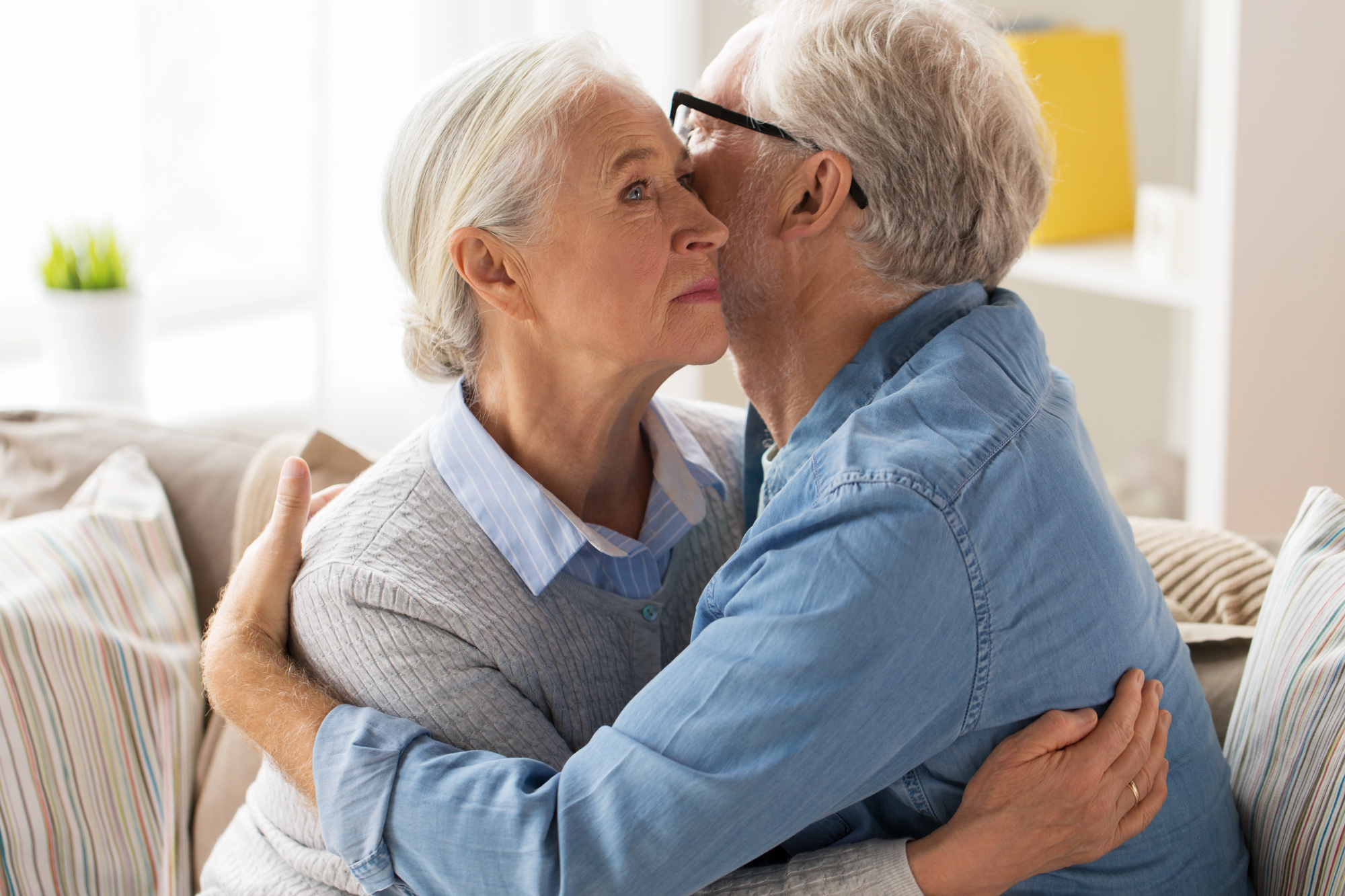 An elderly couple with gray hair is sitting on a couch. The man, wearing glasses and a blue shirt, is embracing the woman, who is wearing a gray sweater and a collared shirt. They are surrounded by soft striped pillows.