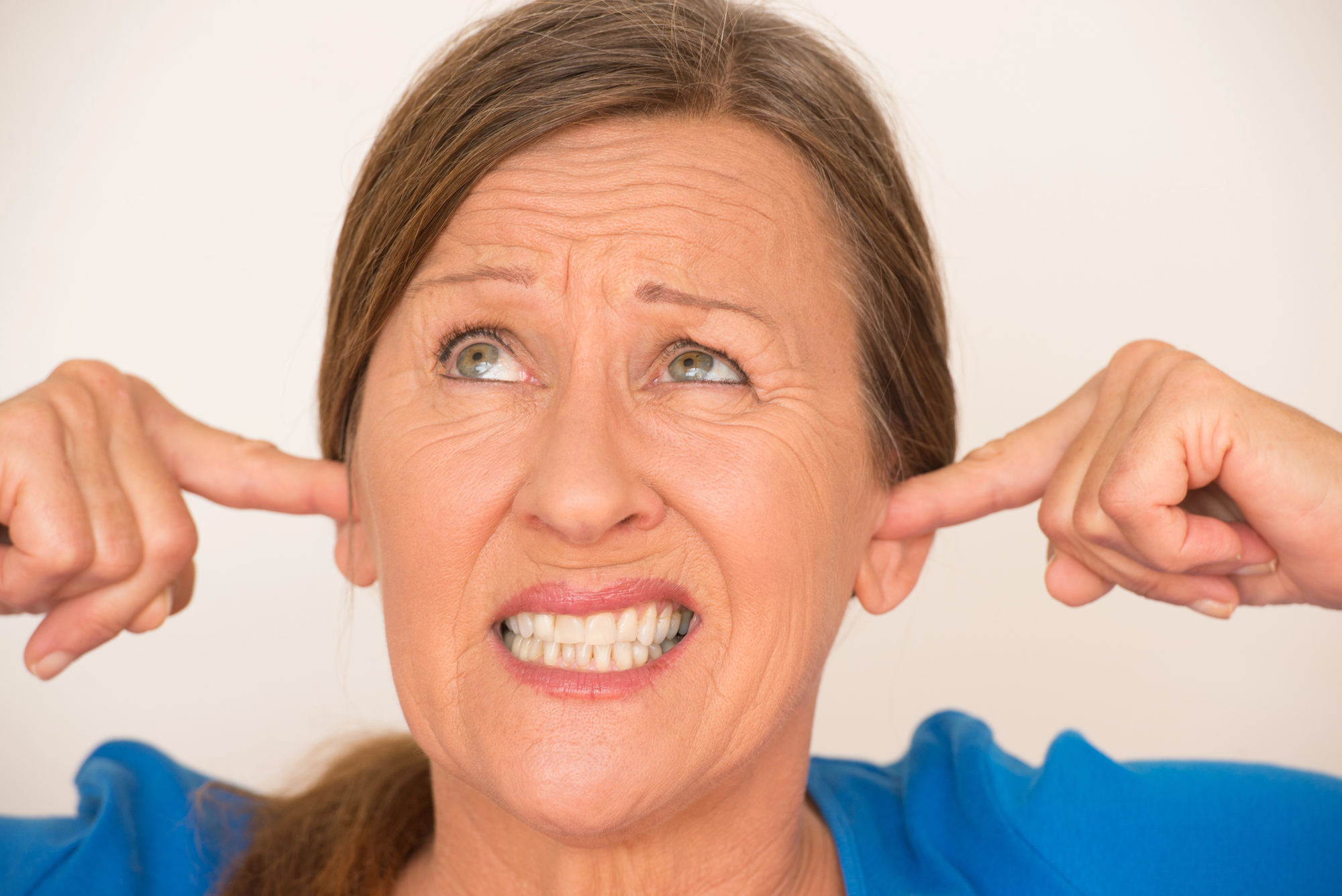 A woman with a worried expression covers her ears with her fingers. She is wearing a blue top and looking upwards, with her mouth slightly open.