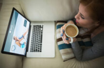 A woman, seated on a couch, holds a coffee mug and looks at a laptop displaying an online dating website with a happy couple on the screen. A striped pillow is beside her.