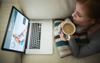 A woman, seated on a couch, holds a coffee mug and looks at a laptop displaying an online dating website with a happy couple on the screen. A striped pillow is beside her.