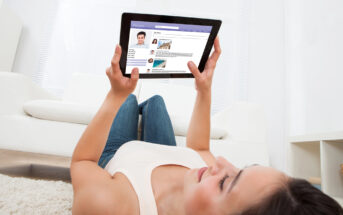 A woman lying on a carpet, holding a tablet displaying a social media profile, with a modern white sofa and blinds in the background. She appears relaxed and focused on the screen.