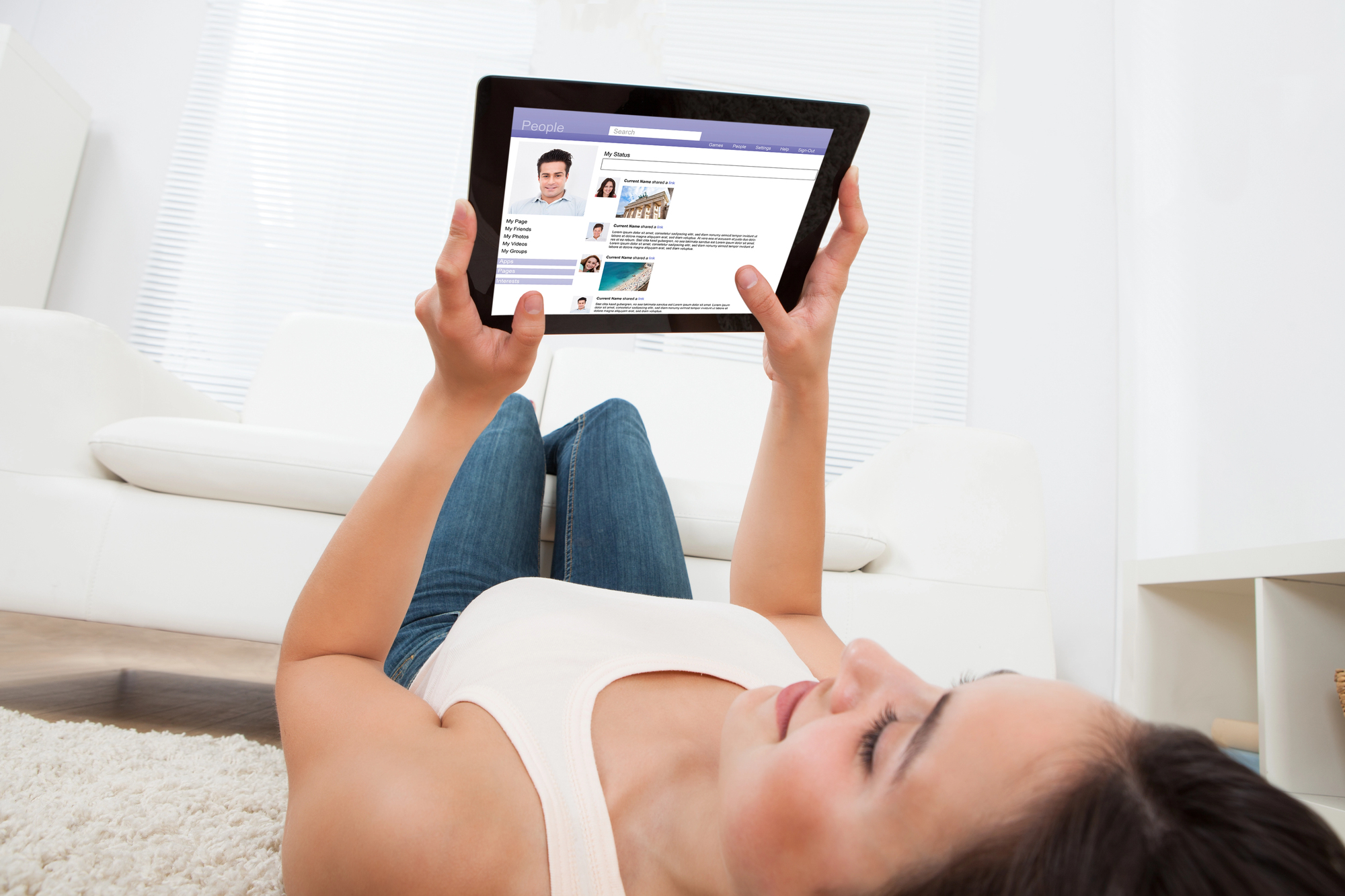 A woman lying on a carpet, holding a tablet displaying a social media profile, with a modern white sofa and blinds in the background. She appears relaxed and focused on the screen.