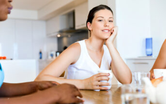 A woman smiles while sitting at a kitchen table, resting her head on her hand and holding a cup. Two other blurred figures are present, suggesting a casual gathering. The background shows a modern, bright kitchen.