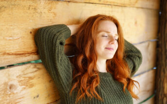 A person with long red hair wearing a green sweater is leaning back against a wooden wall with eyes closed, appearing relaxed and content.