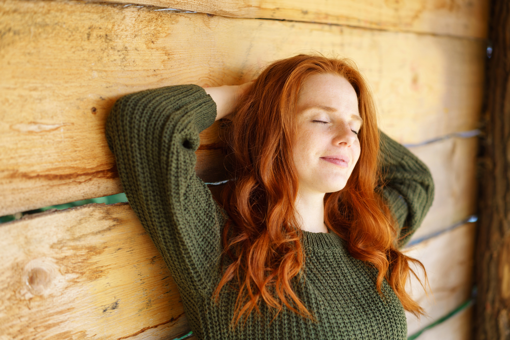 A person with long red hair wearing a green sweater is leaning back against a wooden wall with eyes closed, appearing relaxed and content.