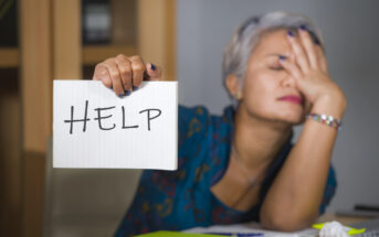 A person with short gray hair is sitting at a table, holding a sign that says "HELP." They appear stressed, with their other hand covering their face. There are crumpled papers and colorful folders on the table.