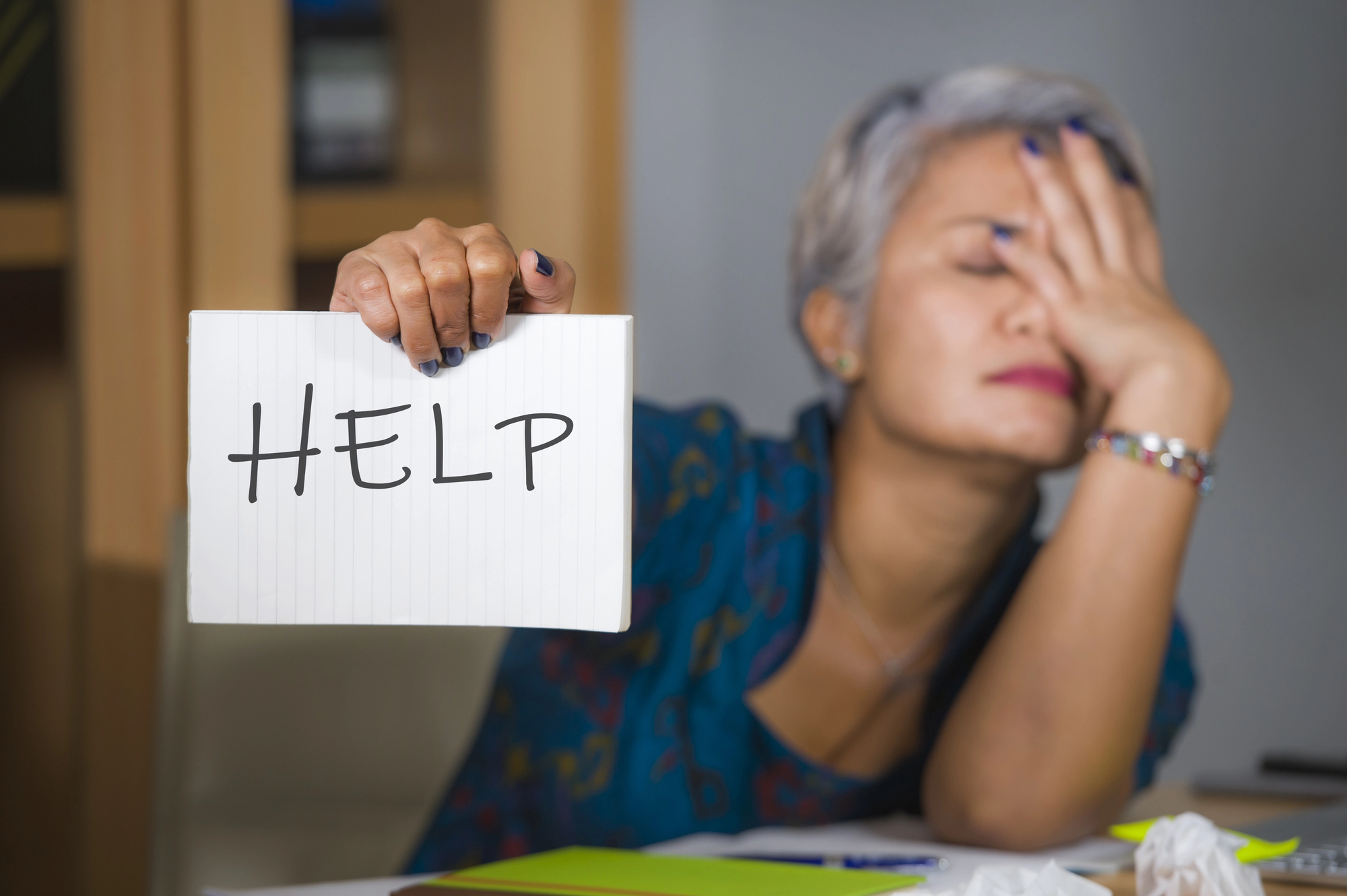 A person with short gray hair is sitting at a table, holding a sign that says "HELP." They appear stressed, with their other hand covering their face. There are crumpled papers and colorful folders on the table.