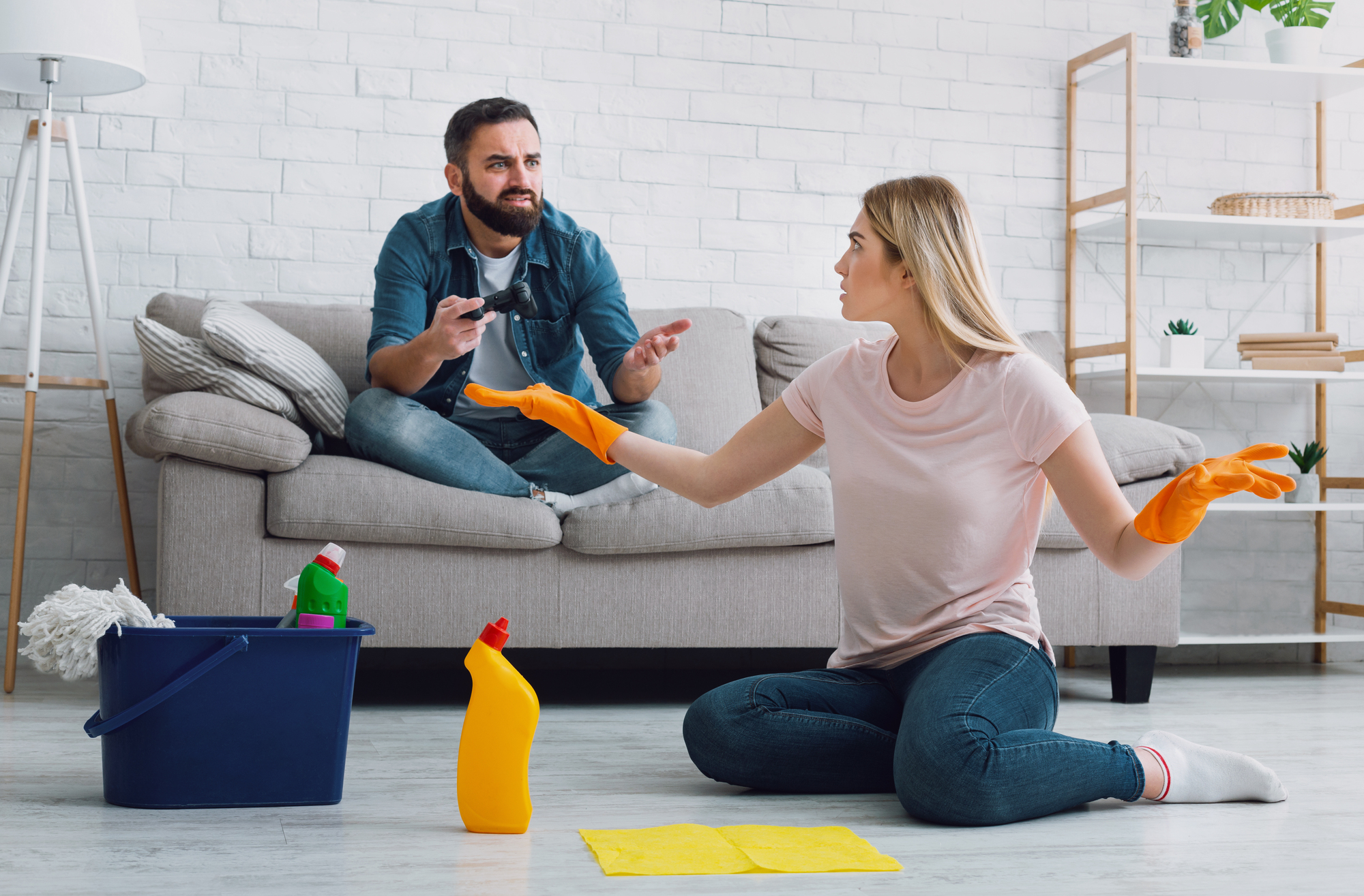 A woman wearing orange gloves sits on the floor with cleaning supplies, gesturing towards a man sitting on a couch holding a TV remote. The room features a modern decor with a plant and a bright, tidy setting.