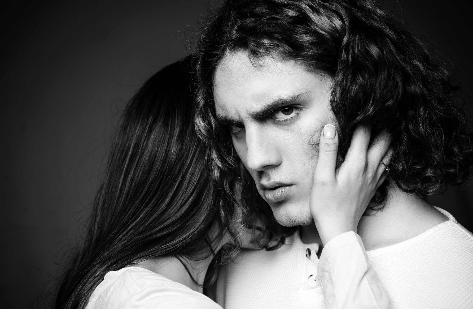 Black and white photo of a person with long curly hair, looking intensely at the camera. Another person, partially visible, holds their face with one hand, creating a dramatic, intimate atmosphere.