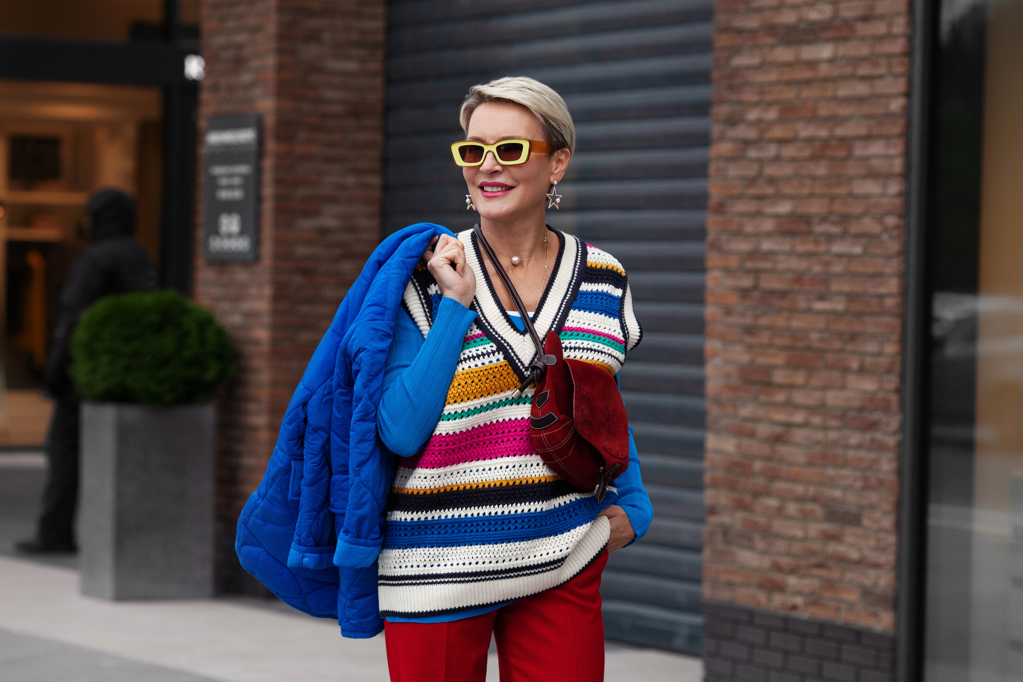 A stylish person wearing yellow sunglasses, a striped sweater vest, blue shirt, and red pants holds a blue jacket over their shoulder and carries a red bag. They are standing outside near a brick wall and a building entrance.