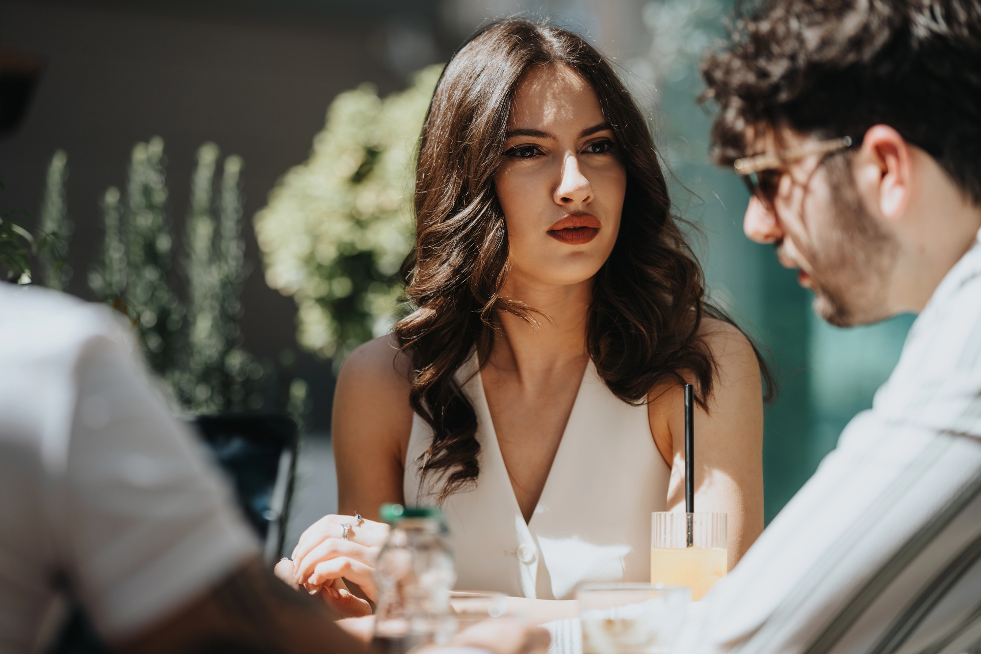 A woman with long brown hair sits at an outdoor table, wearing a white sleeveless top. She looks intently across the table at a person wearing sunglasses. Sunlight filters through greenery in the background.