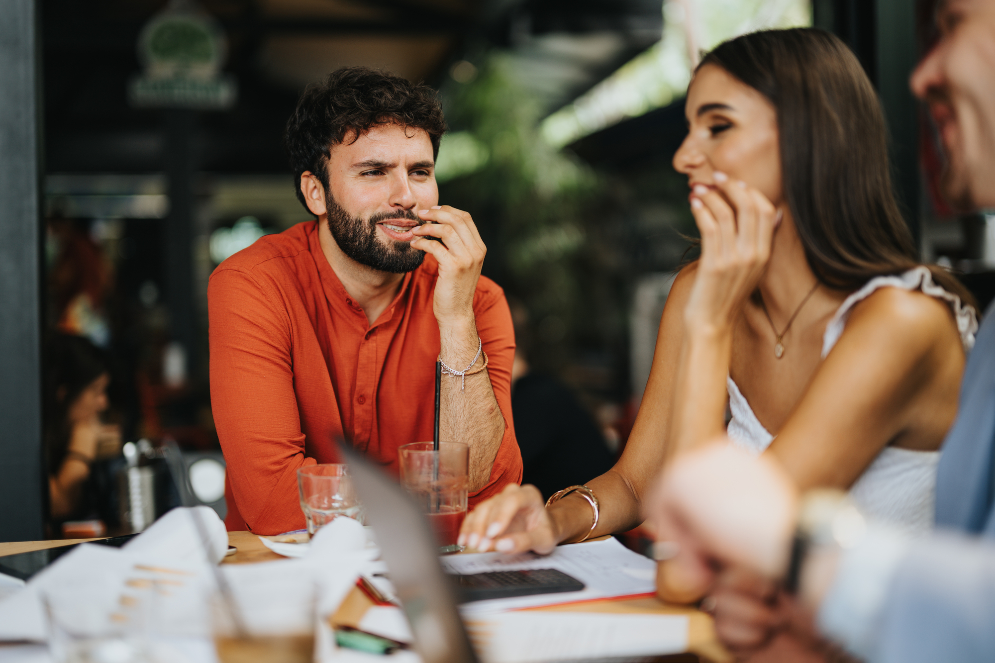 A man and a woman sit at a table, engaged in lively conversation. The man wears an orange shirt and has a thoughtful expression. The woman, in a white top, smiles. Papers and drinks are on the table, suggesting a casual meeting.