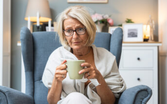 Elderly woman with short blonde hair and glasses sitting in a blue armchair, holding a green mug. She's wearing a light shawl and appears relaxed in a cozy room with soft lighting, decorated with photos and candles.