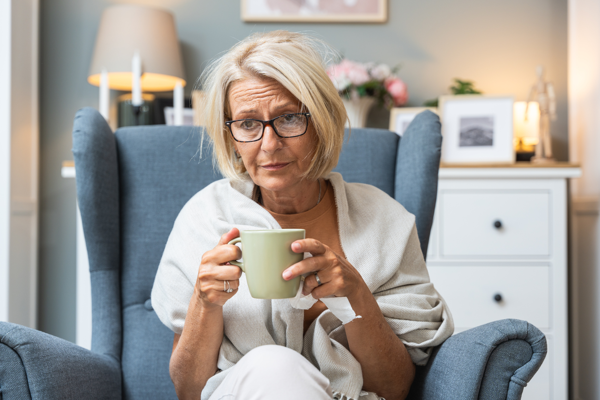 Elderly woman with short blonde hair and glasses sitting in a blue armchair, holding a green mug. She's wearing a light shawl and appears relaxed in a cozy room with soft lighting, decorated with photos and candles.