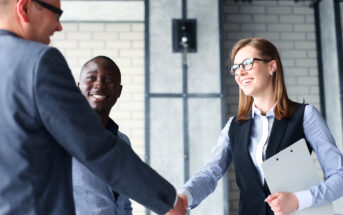 Three people in a business setting smile and shake hands. Two men and one woman, all in professional attire, stand in an office with brick walls and industrial decor. The woman holds a clipboard.