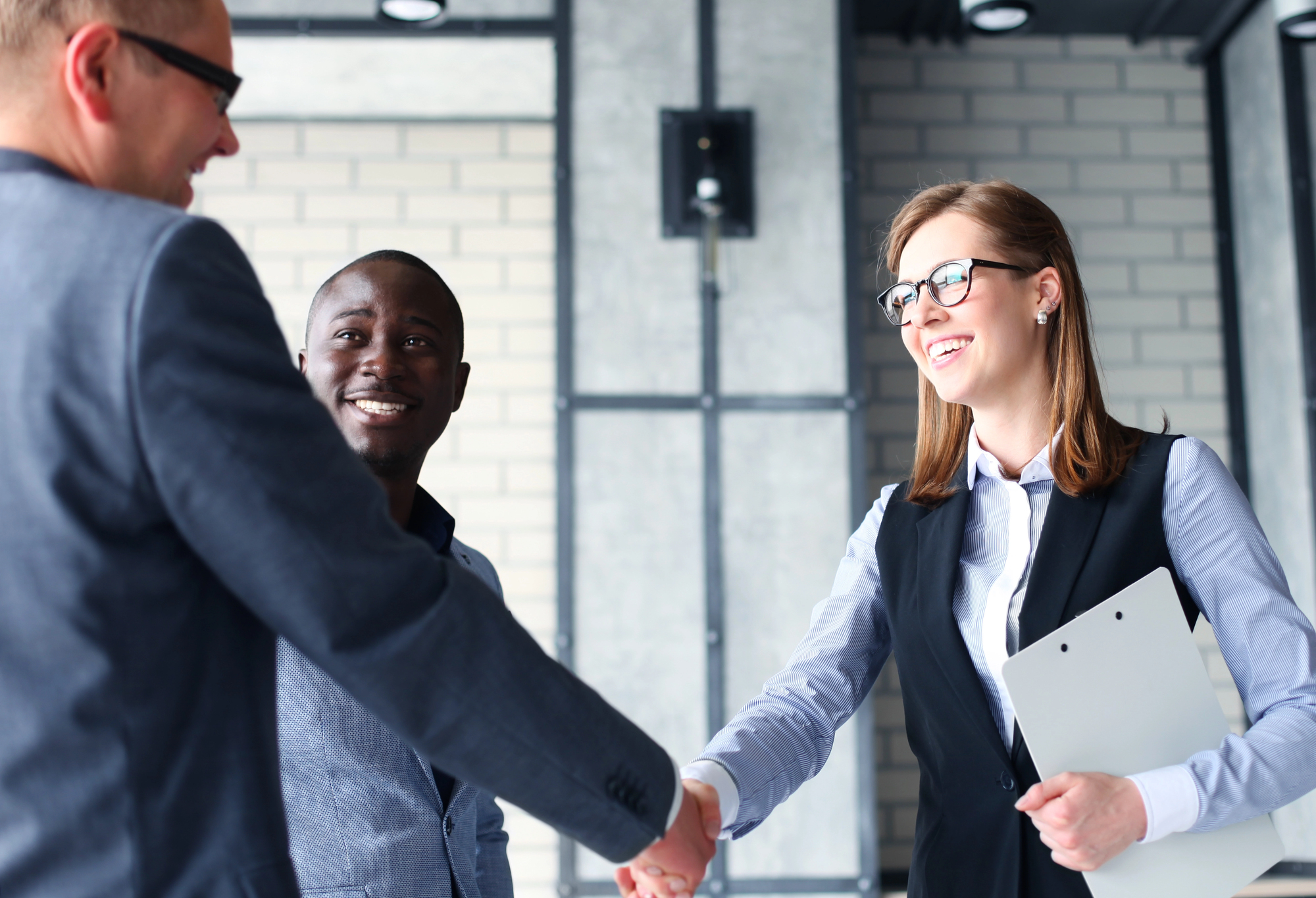 Three people in a business setting smile and shake hands. Two men and one woman, all in professional attire, stand in an office with brick walls and industrial decor. The woman holds a clipboard.