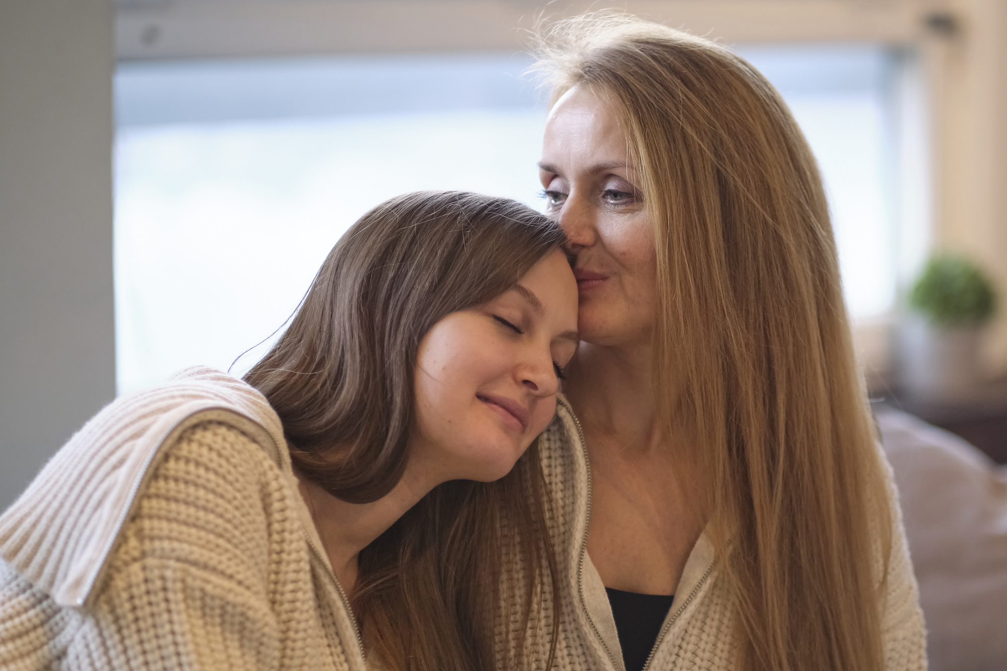 A woman gently kisses the forehead of a girl resting her head on her shoulder. Both are wearing cozy sweaters and appear content. The background is softly blurred, giving a peaceful, intimate atmosphere.