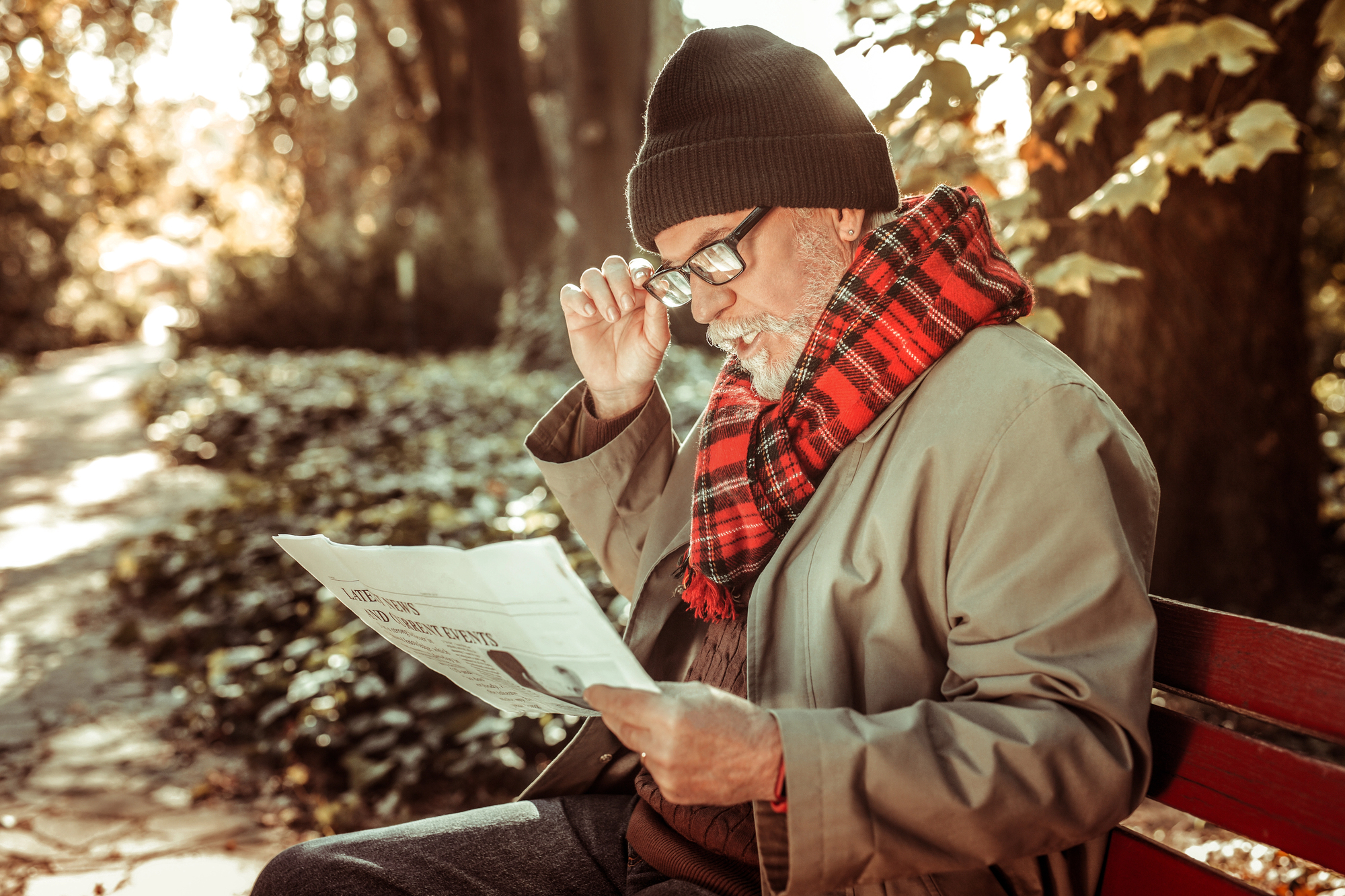 An elderly man wearing a beanie, glasses, and a red plaid scarf sits on a park bench. He is reading a newspaper surrounded by autumn trees and fallen leaves. The sunlight filters through the branches, creating a warm atmosphere.