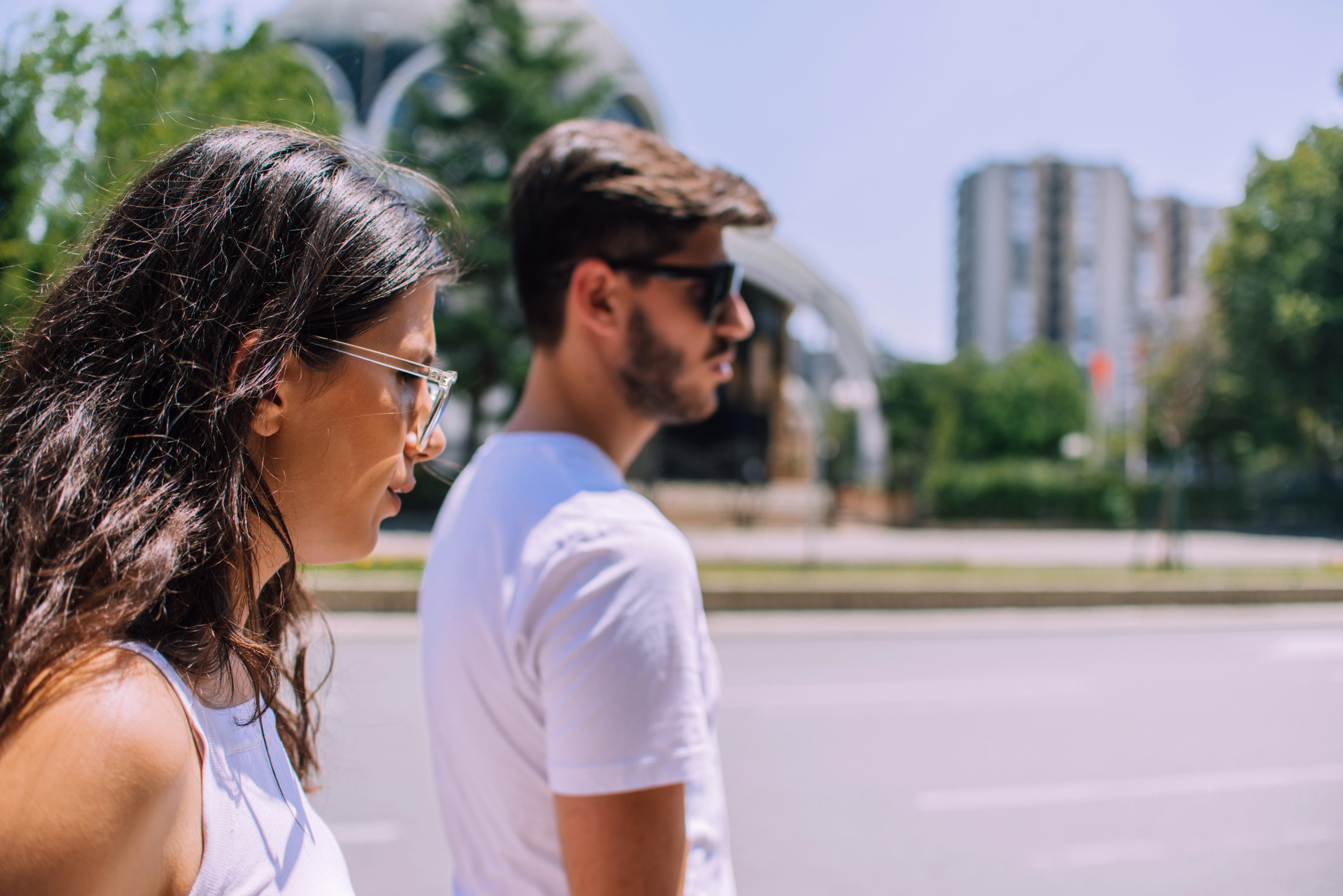 A woman and a man, both wearing sunglasses and white shirts, walk side by side on a sunny day. They are on a sidewalk with blurred buildings and trees in the background.