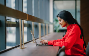 A person in a red hoodie works on a laptop at a long wooden desk by large windows in a modern building. Natural light floods the space, and the view outside is slightly blurred, suggesting an urban setting.