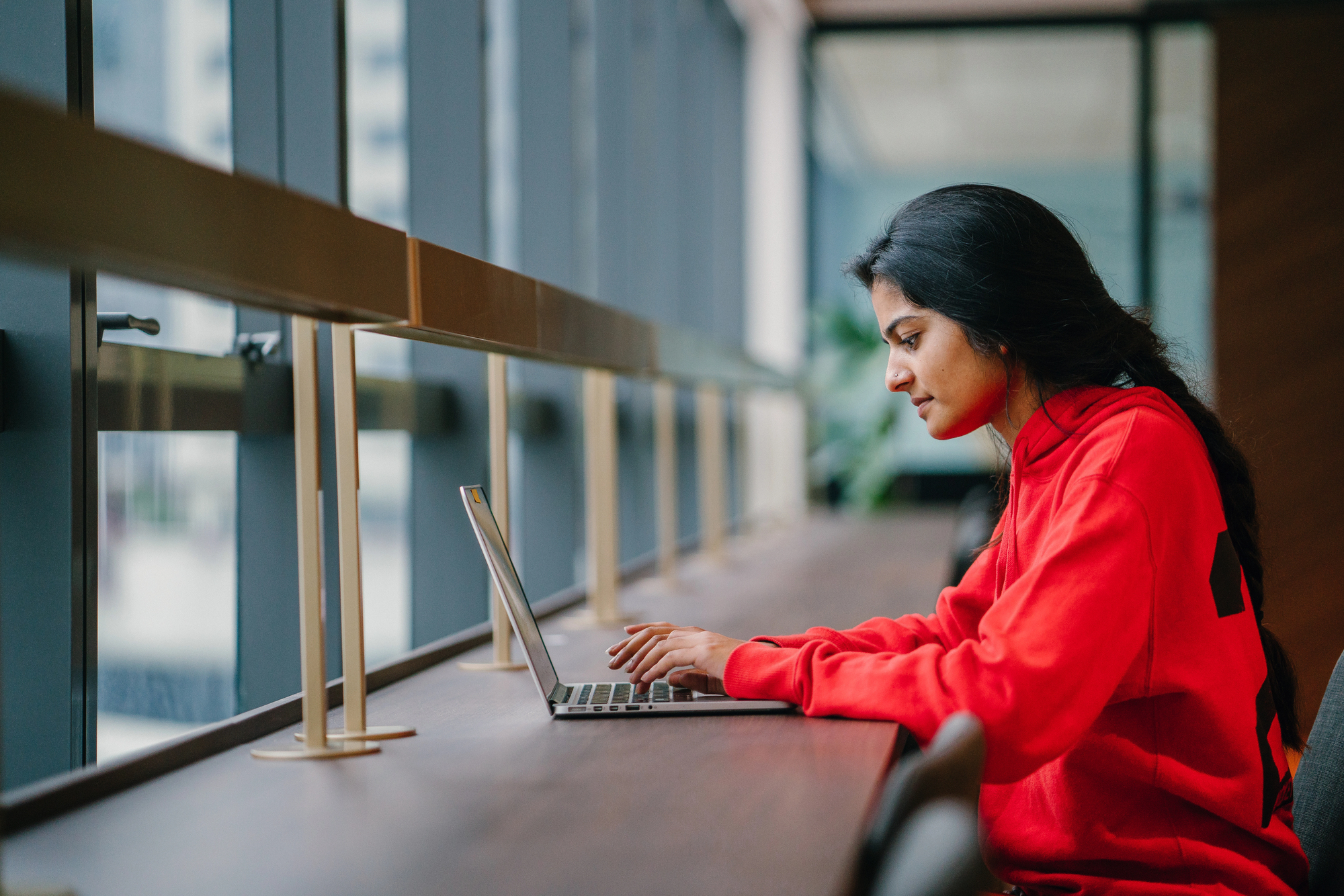 A person in a red hoodie works on a laptop at a long wooden desk by large windows in a modern building. Natural light floods the space, and the view outside is slightly blurred, suggesting an urban setting.