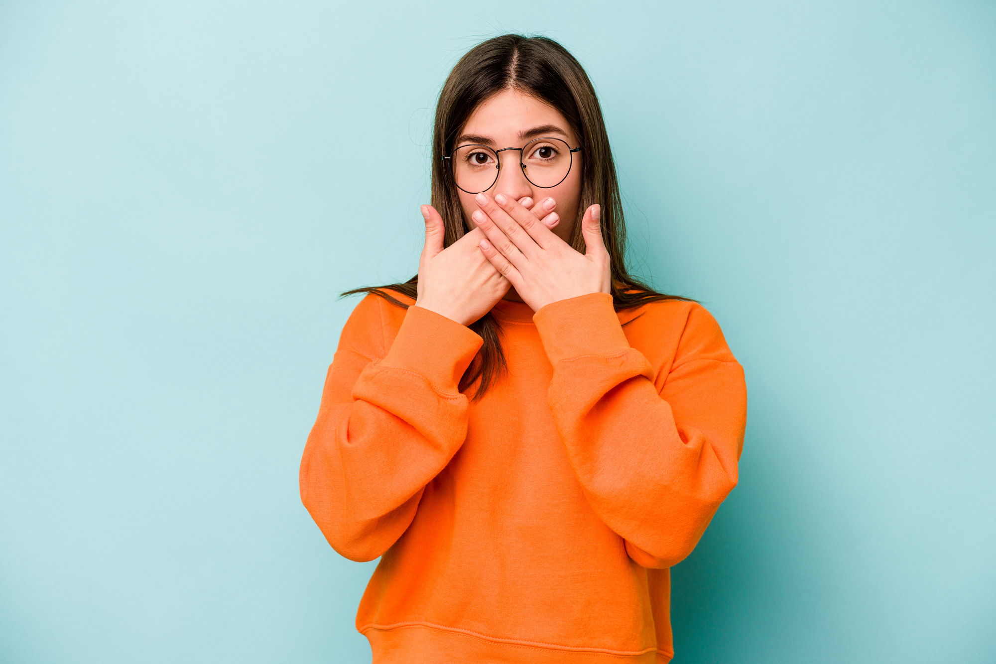 A person wearing glasses and an orange sweatshirt covers their mouth with both hands, standing against a light blue background. They have a surprised or shocked expression.