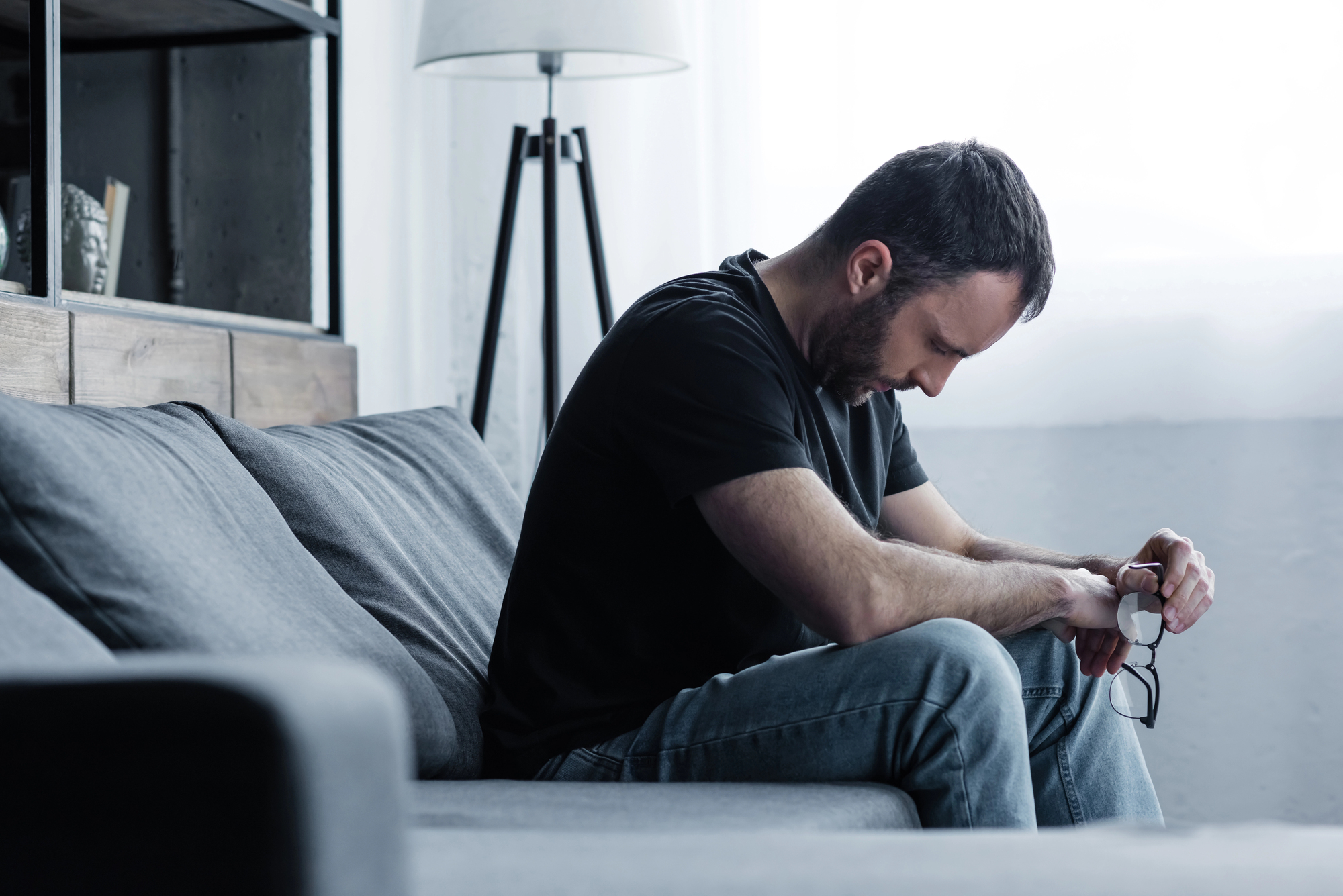 A man sits on a gray couch, looking down intently at his lap while holding a pair of glasses. He is wearing a black t-shirt and jeans. A floor lamp stands in the background, near a bright window.