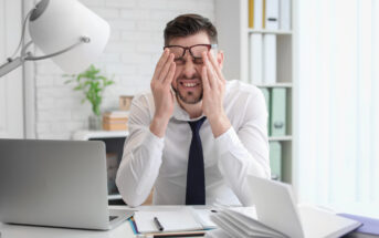 A man in a white shirt and tie sits at a desk with a laptop, holding his glasses and rubbing his eyes in frustration or fatigue. Papers and a desk lamp are visible, with a bright window in the background.