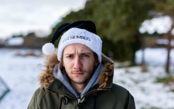 A person wearing a black and white Santa hat with "BAH HUMBUG" written on it stands outdoors on a snowy day. They wear a green parka with a fur-lined hood and look directly at the camera. Trees and snow-covered ground are in the background.