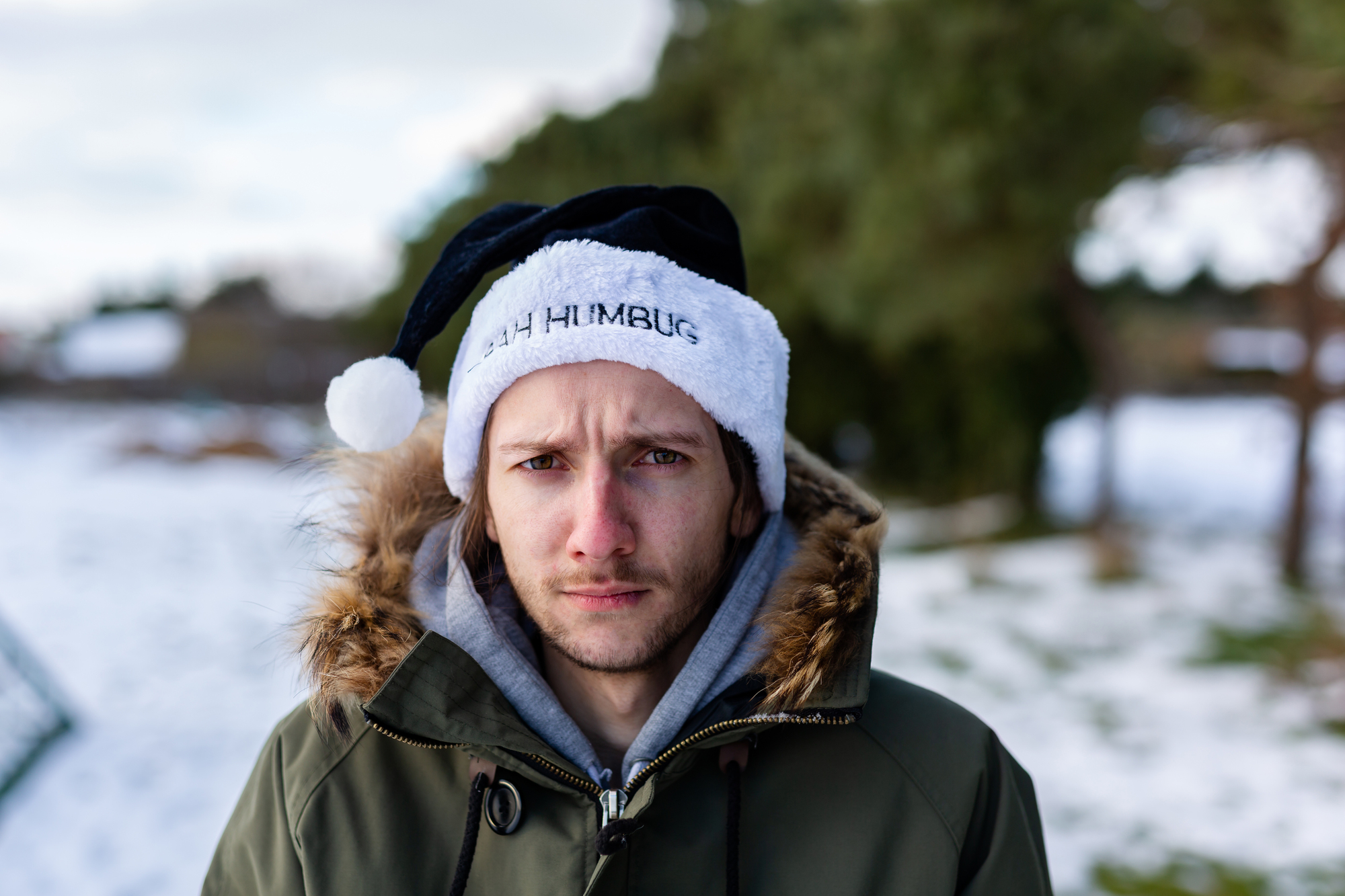 A person wearing a black and white Santa hat with "BAH HUMBUG" written on it stands outdoors on a snowy day. They wear a green parka with a fur-lined hood and look directly at the camera. Trees and snow-covered ground are in the background.