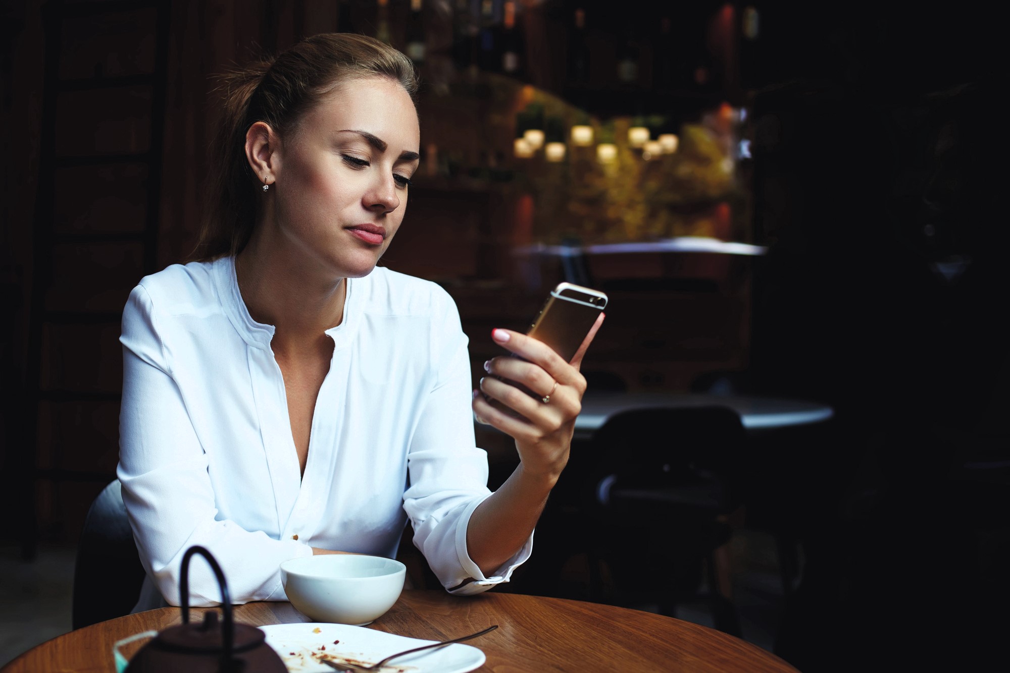 A woman in a white blouse sits at a wooden table in a dimly lit cafe, looking at her smartphone. There is a white bowl and a teapot on the table in front of her. The background features blurred lights and dark furniture.