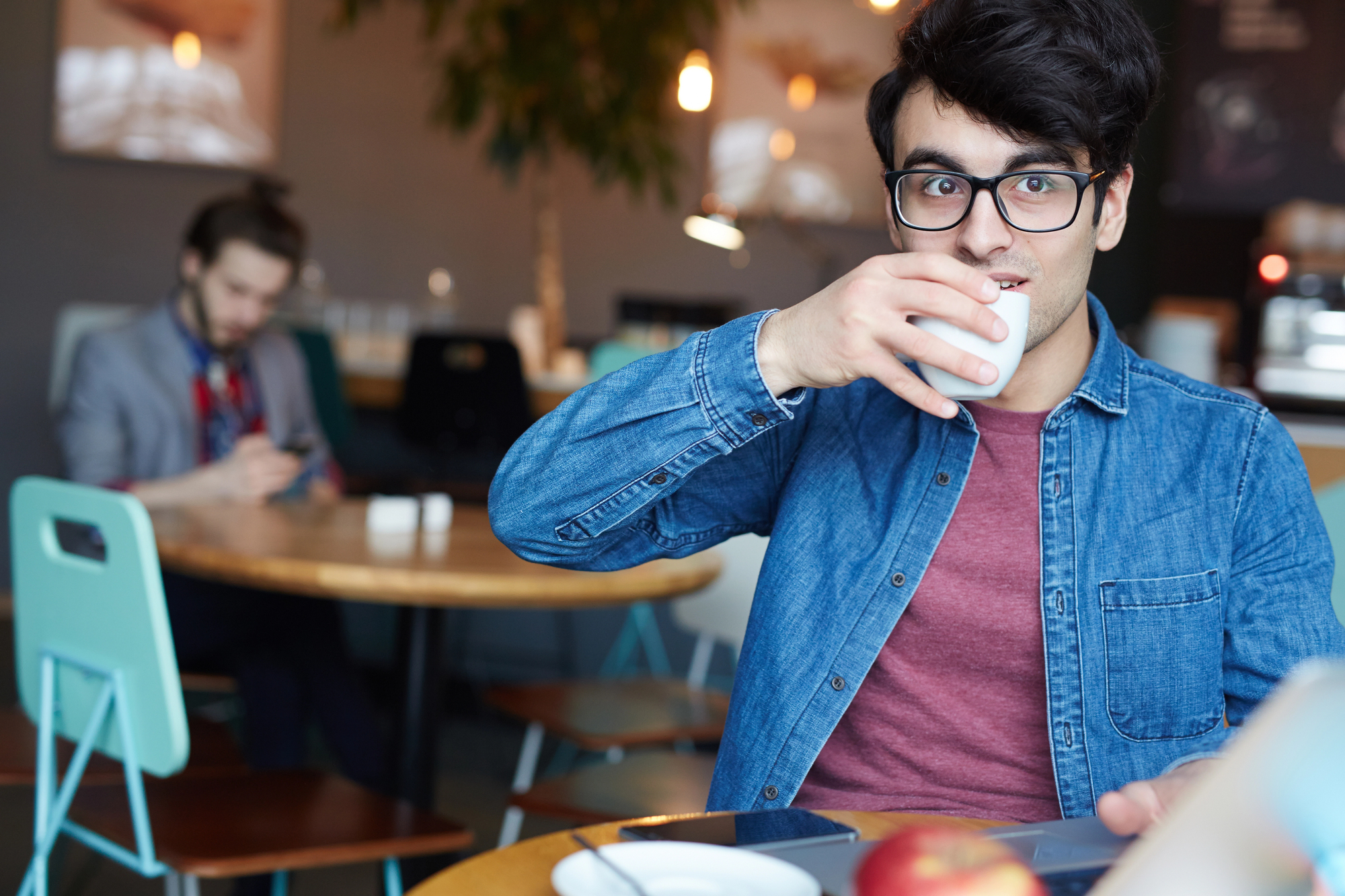 A young man with glasses and dark hair drinks coffee in a cafe, wearing a denim shirt and red t-shirt. He's seated at a table with a smartphone and an apple. In the background, another person is using a phone.