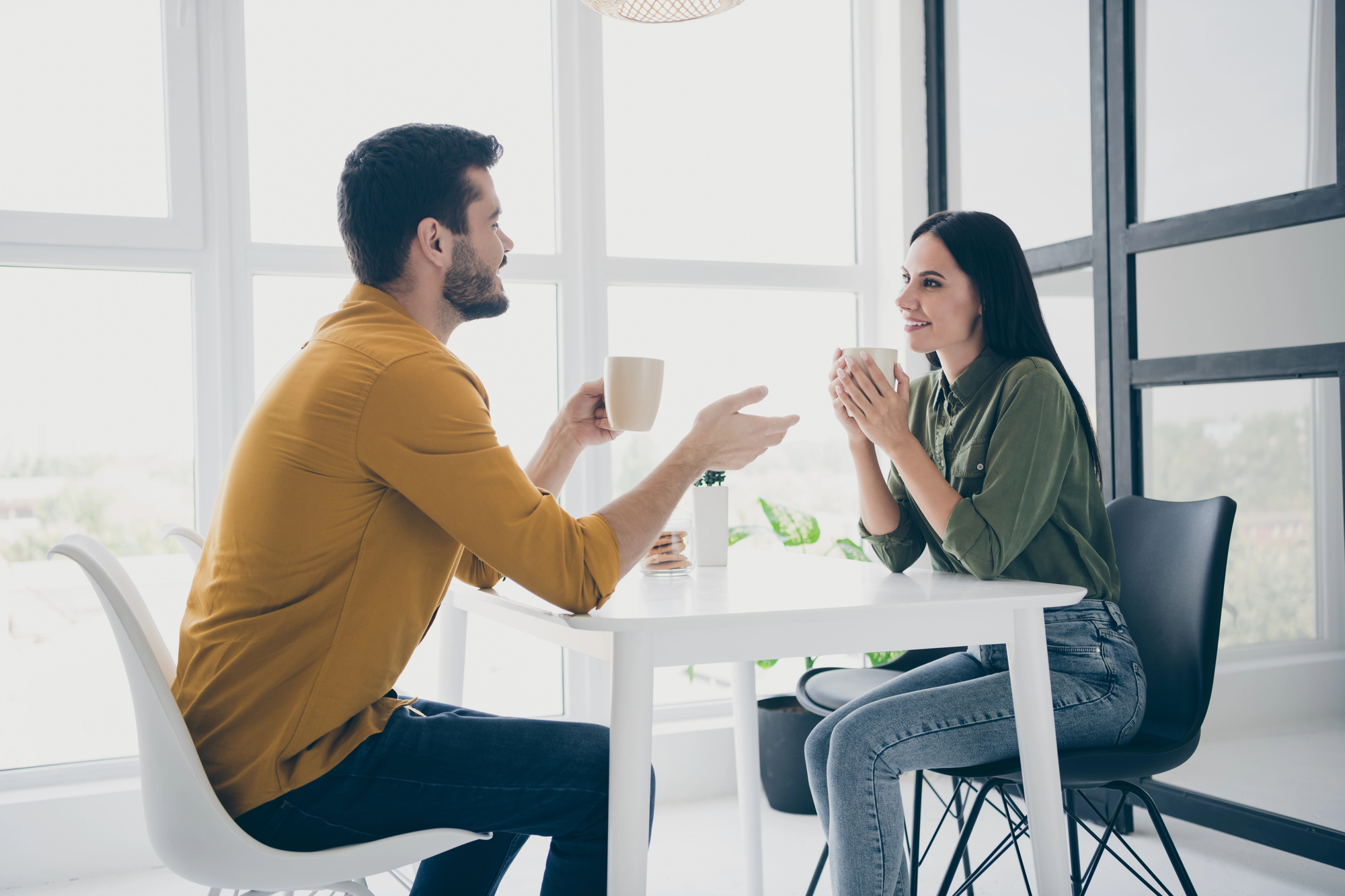 Two people are sitting at a table in a bright room, holding coffee mugs and smiling at each other. One wears a yellow shirt and jeans, the other a green shirt and jeans. There are large windows in the background.