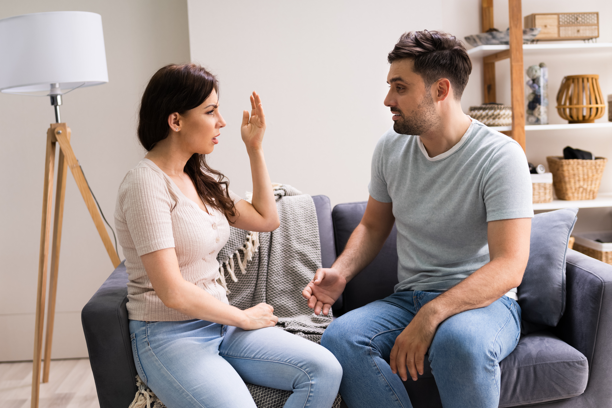 A woman and a man are sitting on a couch, engaged in a serious conversation. The woman is gesturing with her hand raised, while the man looks at her attentively. They are in a cozy living room with shelves and a lamp in the background.